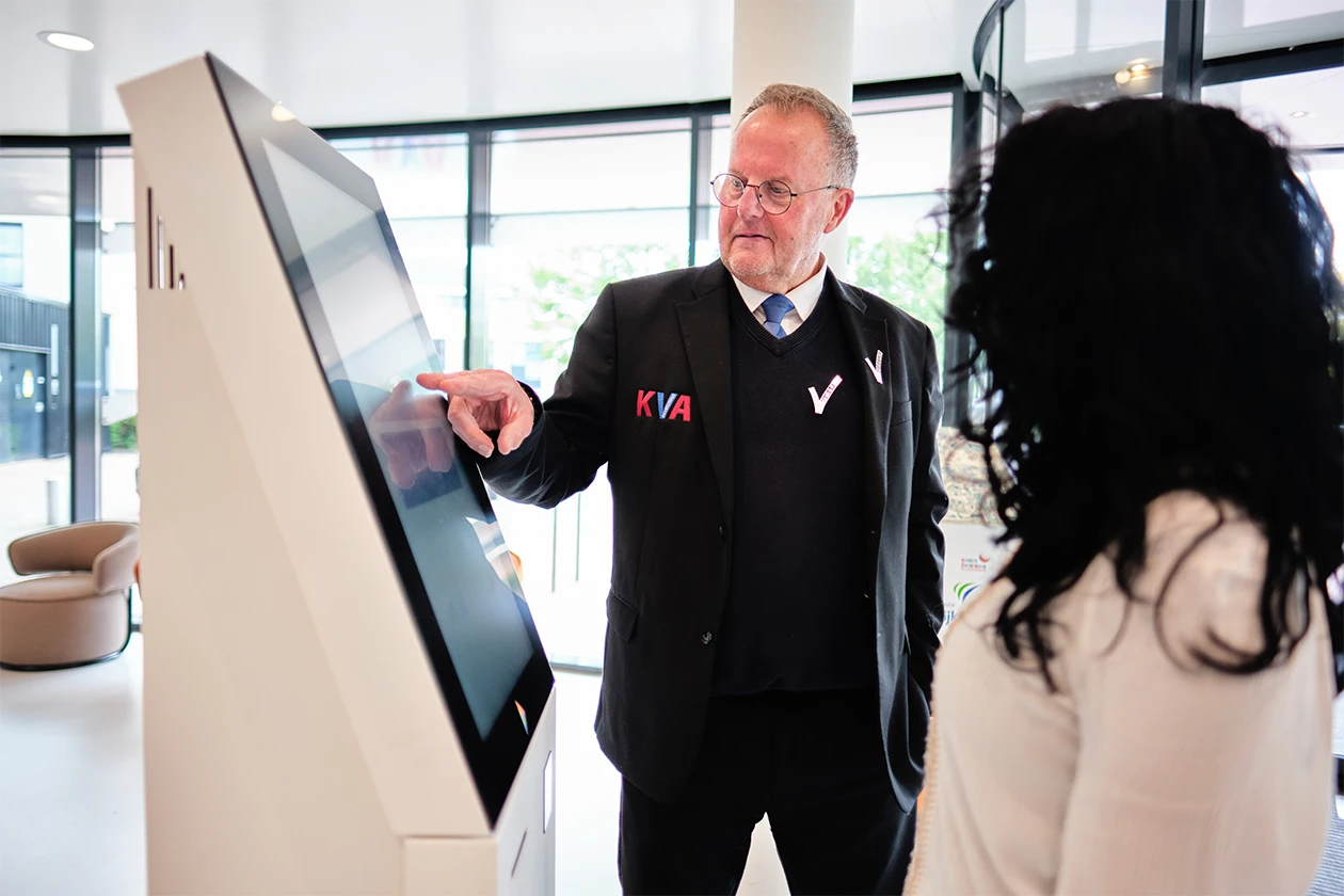 Medewerker van KVA Beveiliging staat in uniform met een bezoeker bij een aanmeldscherm van een receptie